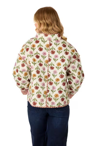 A woman viewed from the back wearing a cream-colored jacket with a dense multicolor floral pattern in pink, red, orange, and green, paired with dark blue jeans, against a plain white background.