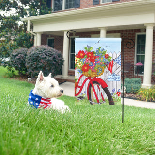 Dog sitting on grass with a decorative garden flag featuring a bicycle and flowers in front of a house.