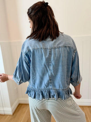 Person wearing a denim jacket with ruffled hem in a room with white walls and wooden floor.