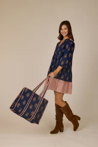 A woman modeling a navy blue patterned tunic dress with a pink and white striped hem, holding a matching navy tote bag with pink and white trim, set against a solid beige studio background.
