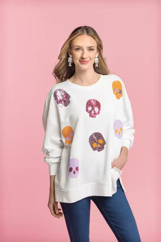 A woman modeling a white crewneck sweatshirt decorated with colorful skull prints in purple, orange, and pink, paired with blue jeans against a solid pink background.