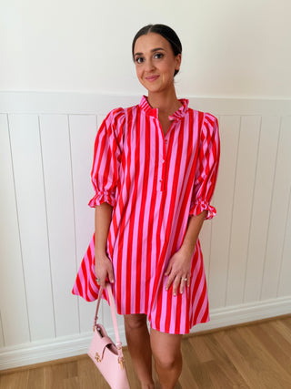 Woman wearing a red and pink striped dress holding a pink handbag against a white paneled wall.