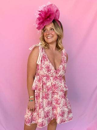 Woman wearing a pink floral dress and matching fascinator against a pink background