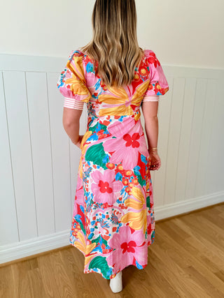 Woman wearing a colorful floral dress standing against a white paneled wall.