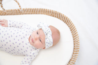 A baby lying on their side in a woven oval bassinet, wearing a white long-sleeve bodysuit with a small purple pattern and a matching white bow headband.