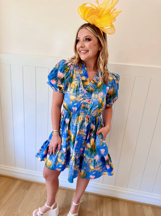 Woman wearing a blue floral dress with a yellow fascinator against a white paneled wall.