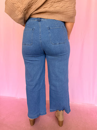 A person shown from the back wearing light-to-medium blue wide-leg cropped jeans, a beige cable-knit sweater, and beige heels against a pink backdrop.