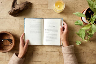 Person reading an open book on a wooden table with a candle and plant