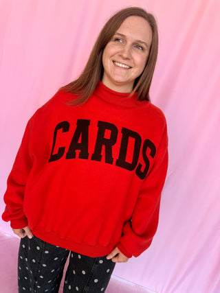 A smiling woman with long brown hair wears a bright red mock-neck sweatshirt with the word 'CARDS' in large black block letters across the chest, paired with black pants with white polka dots, set against a pink draped fabric backdrop.