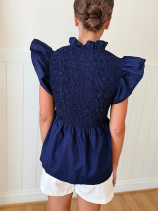 Person wearing a navy smocked top with ruffled sleeves against a white wall.
