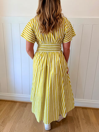 Woman wearing a yellow and white striped dress against a white paneled wall.