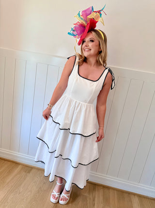 Woman wearing a white dress with black trim and a colorful headpiece against a white paneled wall.