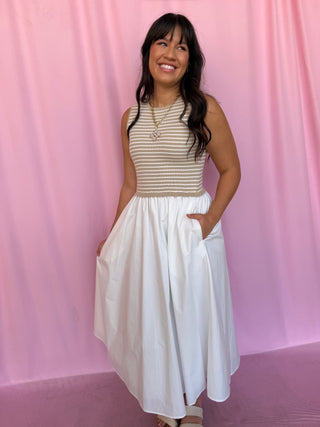 A woman modeling a sleeveless dress with a beige and white striped bodice and a flowing white skirt with side pockets, standing against a pink curtain backdrop.