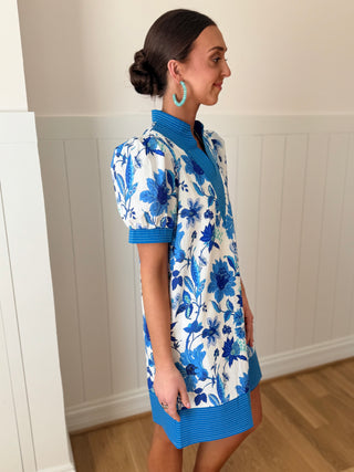 Woman wearing a blue and white floral dress with a matching cardigan in a room with wooden flooring and white walls.
