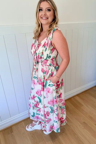 Woman wearing a floral dress and pink hat indoors against a white paneled wall.
