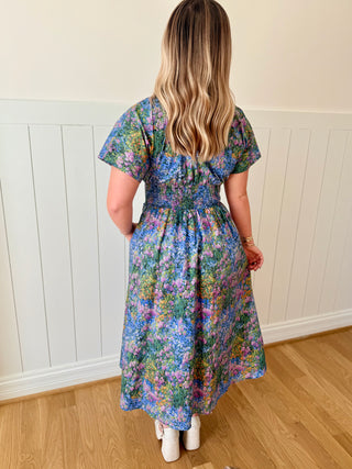 Woman wearing a floral dress standing in a room with wooden flooring and white walls.