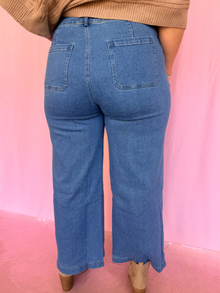 Rear view of a person wearing blue wide-leg cropped jeans with a tan ribbed sweater and tan boots, standing against a pink backdrop.