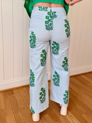 Person wearing white pants with green leaf patterns in a room with wooden flooring and a white paneled wall.