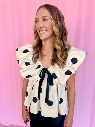 A woman with blonde-highlighted hair smiling, wearing a cream-colored peplum blouse with black polka dots and a ruffled tie neckline, set against a pink backdrop.