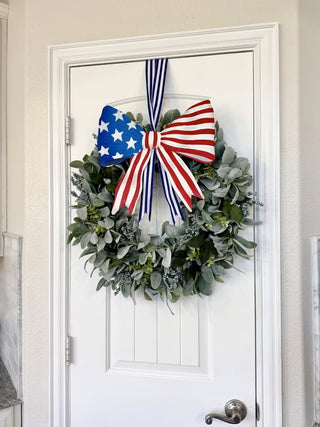 Green wreath with a large red, white, and blue bow on a white door.