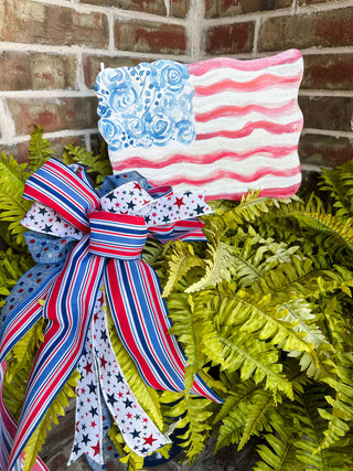 Decorative wreath with a red, white, and blue ribbon and American flag design against a brick wall.