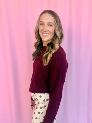 A smiling woman with blonde-highlighted hair stands against a pink draped backdrop, wearing a dark burgundy knit sweater and cream pants with brown polka dots.