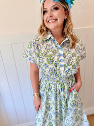 Woman wearing a floral dress with a striped shirt underneath, standing against a white paneled wall.
