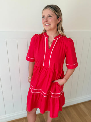 Woman wearing a red dress with white trim against a white paneled wall.