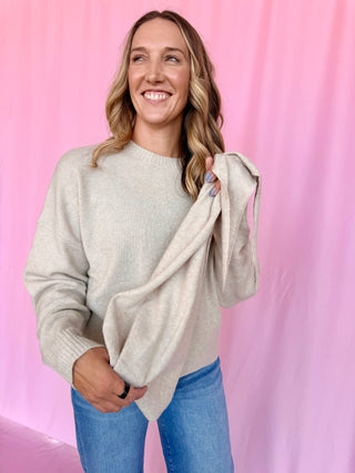 A smiling woman with blonde hair models a light heather gray knit sweater and blue jeans, holding up the sleeve of the sweater against a pink fabric backdrop.