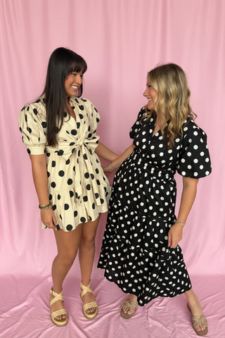 Two women wearing polka dot dresses standing against a pink background