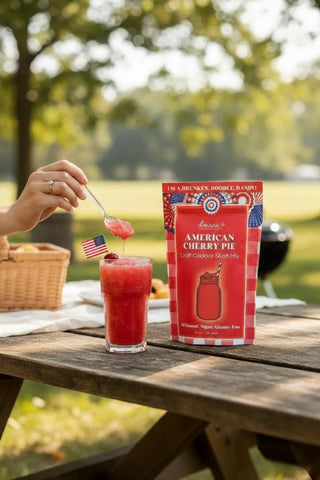 Person stirring a red drink in a glass with a package of American Cherry Pie mix on a wooden table outdoors.