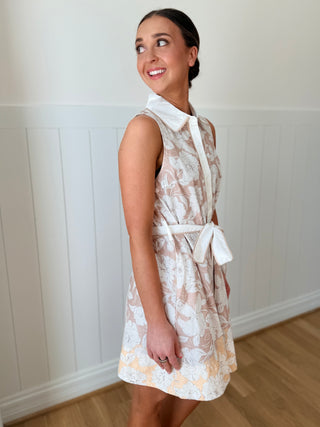 Woman wearing a floral dress with a white apron against a white paneled wall.
