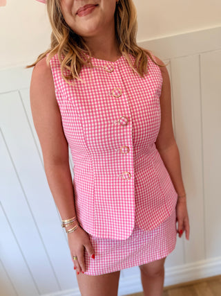 Woman wearing a pink gingham dress with a matching jacket against a white paneled wall.
