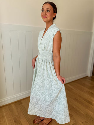 Woman wearing a light green patterned dress standing in a room with wooden flooring and white paneled walls.