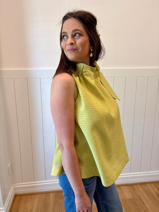 Woman wearing a yellow sleeveless top with a bow detail, standing against a white paneled wall.