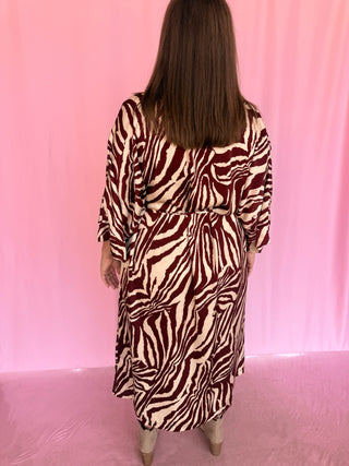 A woman viewed from behind wearing a long-sleeve wrap dress with a bold zebra print in dark red and cream, standing against a pink backdrop.