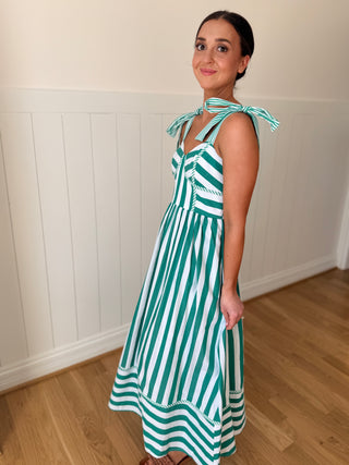 Woman wearing a green and white striped dress with bow straps in a room with wooden flooring and white walls.