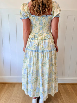 Woman wearing a floral dress with puffed sleeves against a white paneled wall.