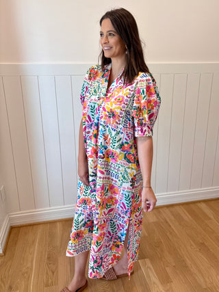 Woman wearing a colorful floral dress standing in a room with wooden flooring and white paneled walls.