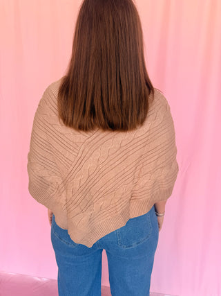 A person viewed from behind wearing a chunky cable-knit cropped sweater in a warm beige/tan color, paired with blue jeans, standing against a pink draped backdrop.