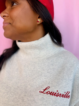 A close-up side profile of a woman wearing a light heather gray turtleneck sweatshirt with 'Louisville' embroidered in red script on the chest, paired with a red cap and a small gold earring, against a pink background.