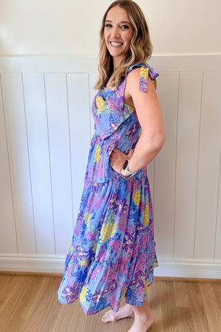 Woman wearing a colorful floral dress against a white paneled wall.