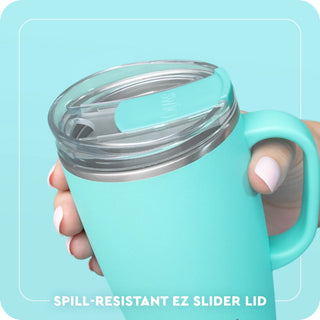 A close-up of a mint green insulated tumbler with a clear lid and a matching mint green spill-resistant EZ slider lid, held by a hand against a light blue background with text at the bottom.