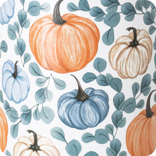 A close-up of a fabric or wallpaper pattern featuring hand-drawn watercolor pumpkins in orange, blue, and cream, surrounded by teal eucalyptus leaves on a white background.