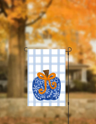 A decorative garden flag hanging on a black metal stand, featuring a blue pumpkin with white floral patterns and an orange bow on a white gingham background. The background is a blurred outdoor autumn scene with orange trees.