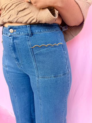 Close-up of a person wearing blue jeans with decorative wavy gold embroidery across the back pocket, paired with a beige ribbed top against a pink background.