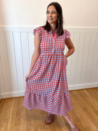 Woman wearing a red and blue checkered dress standing in a room with wooden flooring and white paneled walls.