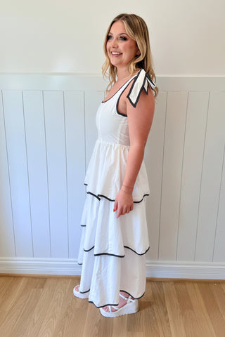 Woman wearing a white dress with black trim and a colorful hat against a white paneled wall.