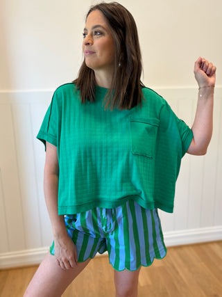 Woman wearing a green oversized top and striped shorts against a white wall.