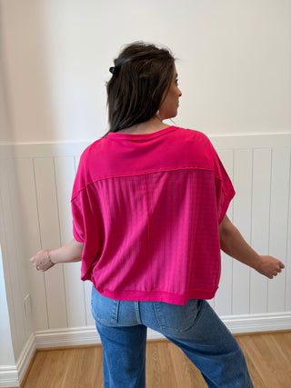 Woman wearing a bright pink top and blue jeans standing in a room with white walls and wooden flooring.